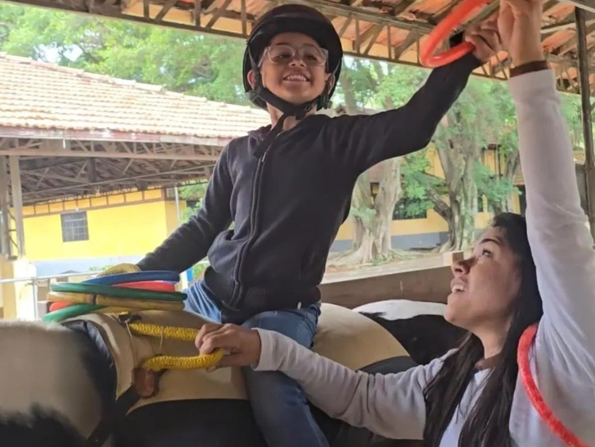 Criança sorridente montando em um touro mecânico, com ajuda de uma mulher